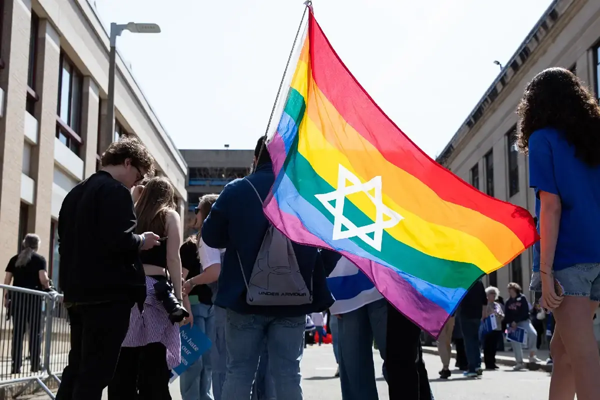 Young adult jewish life boston: Young adults gather outdoors at a community event while holding a rainbow Pride flag featuring the Star of David.