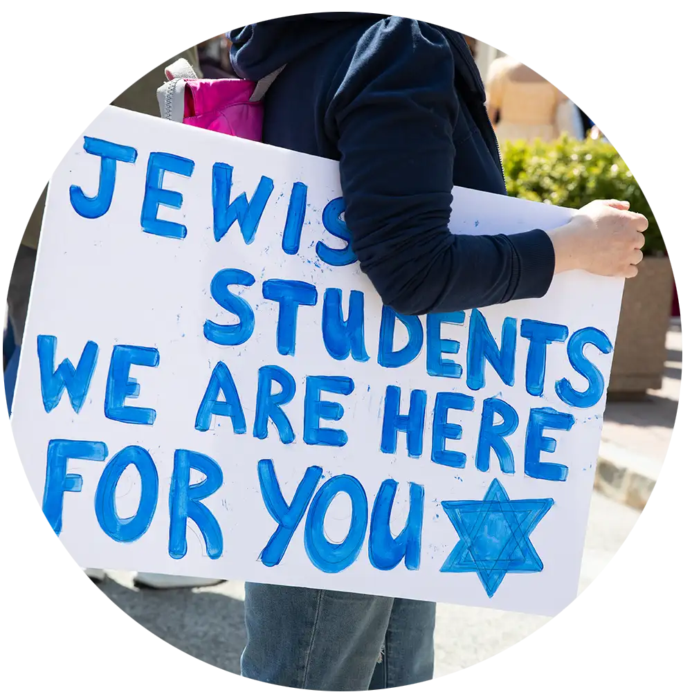 College student holding a sign reading “Jewish students we are here for you,” showing visible support and solidarity on campus