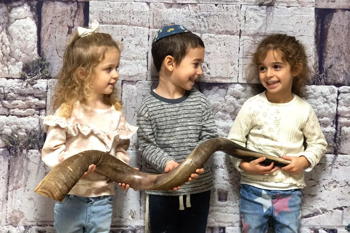 Three young children smiling and holding a shofar at a Jewish family event, highlighting Jewish family support and events for Jewish kids in Boston.